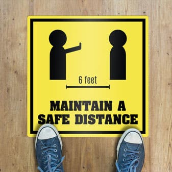 A square floor sticker on a wood floor shows an alert to maintain a safe distance of 6 feet away.