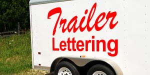 A white trailer with adhesive lettering applied to the side has been parked with grass in the background.  